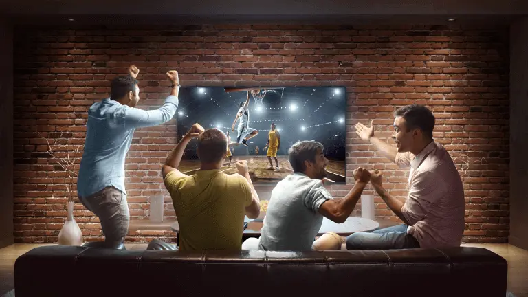 Four friends cheering while watching basketball on a wide screen TV in a living room with theater lighting and surround sound.